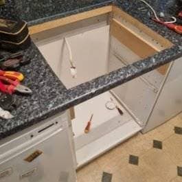 Open kitchen cupboard with tools and wiring exposed, worktop partially removed, and various tools like pliers and screwdrivers scattered nearby, indicating ongoing repair or installation work. Open kitchen cupboard with tools and wiring exposed, worktop partially removed, and various tools like pliers and screwdrivers scattered nearby, indicating ongoing repair or installation work.