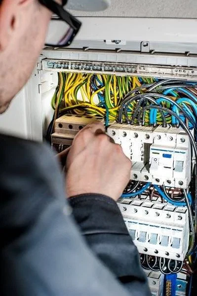 A person wearing glasses and a dark jacket works on an open electrical panel filled with various coloured wires and circuit breakers.