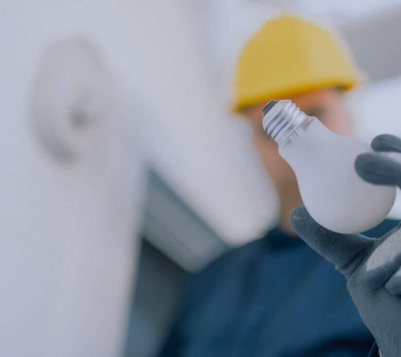 A person wearing a yellow hard hat and gloves holds a frosted light bulb, with the background softly blurred, suggesting they are about to fit or replace it.