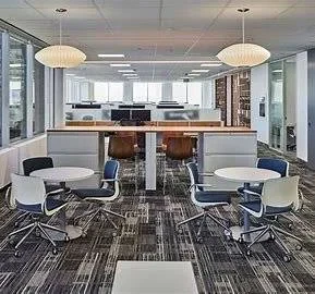 Modern open-plan office with large windows, two round tables with chairs in the foreground, and workstations separated by partitions in the background. Pendant lights hang from the ceiling, creating a bright, welcoming atmosphere. Modern open-plan office with large windows, two round tables with chairs in the foreground, and workstations separated by partitions in the background. Pendant lights hang from the ceiling, creating a bright, welcoming atmosphere.