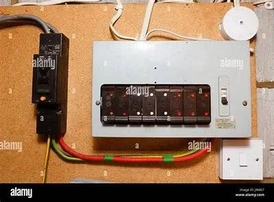 A grey consumer unit with multiple switches is mounted on a wooden wall, with coloured wires connected below and a white socket to the right.