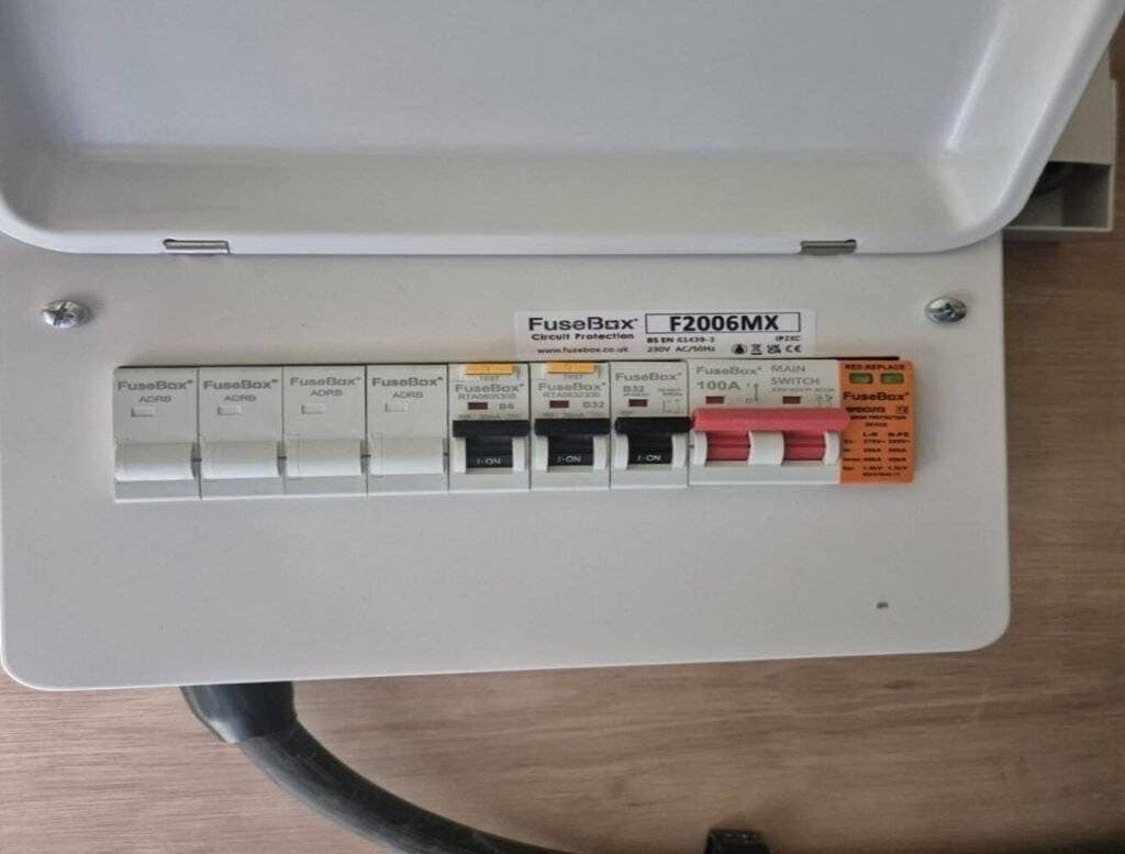 A white consumer unit with labelled switches, including several circuit breakers and a main switch, mounted on a light wood surface. The cover is open, revealing the labelled controls inside.