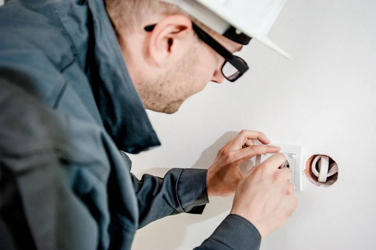A person wearing a hard hat and glasses is installing or repairing an electrical socket on a white wall, using a screwdriver. A person wearing a hard hat and glasses is installing or repairing an electrical socket on a white wall, using a screwdriver.