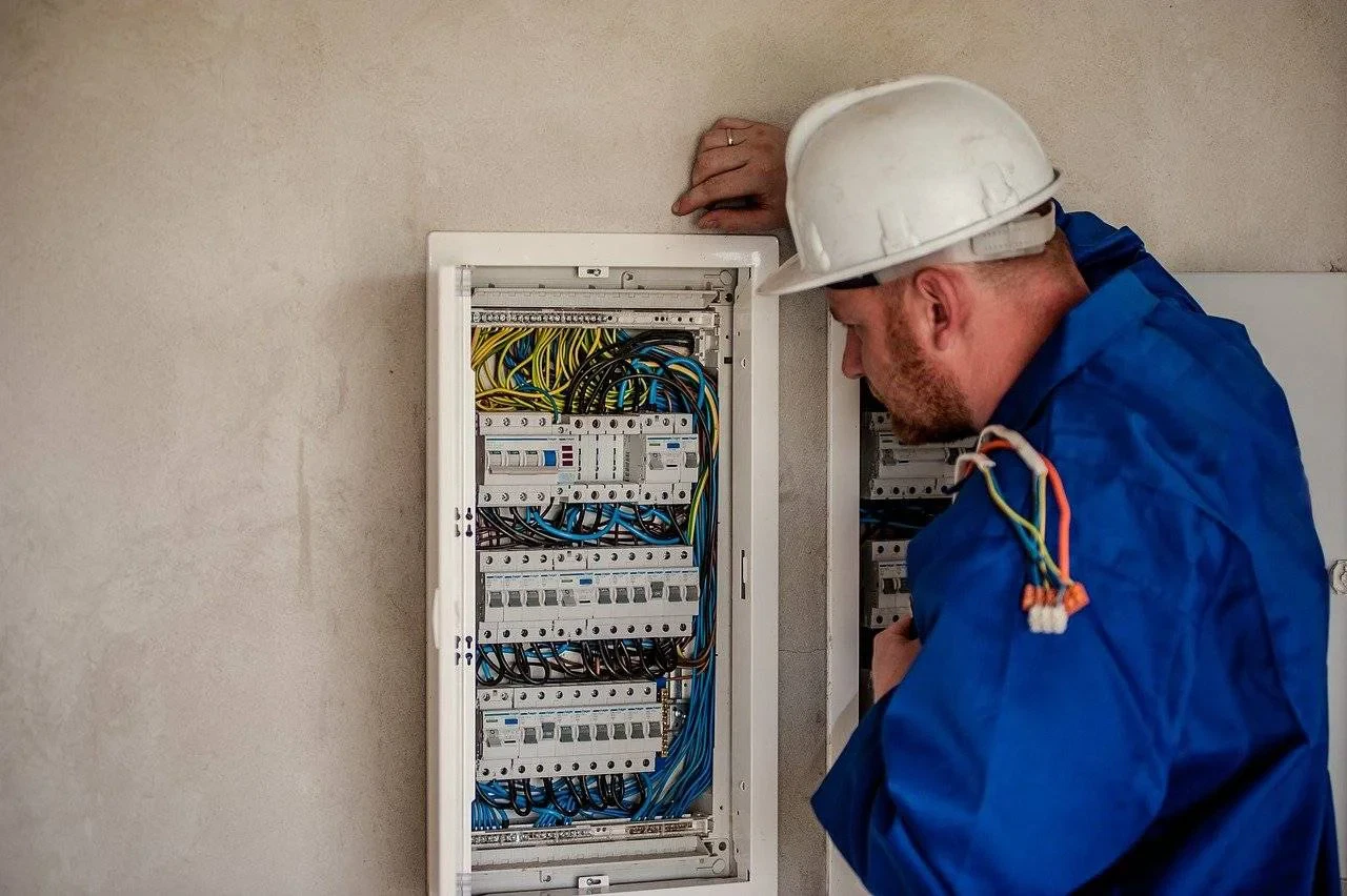 An electrician wearing a white hard hat and blue work clothes is inspecting an open electrical panel with many coloured wires and switches on a beige wall.
