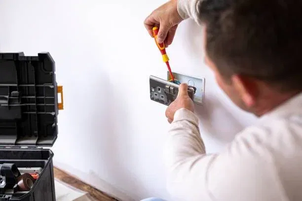 A person uses a screwdriver to install or repair an electrical socket on a white wall, with a toolbox nearby containing various tools and supplies.