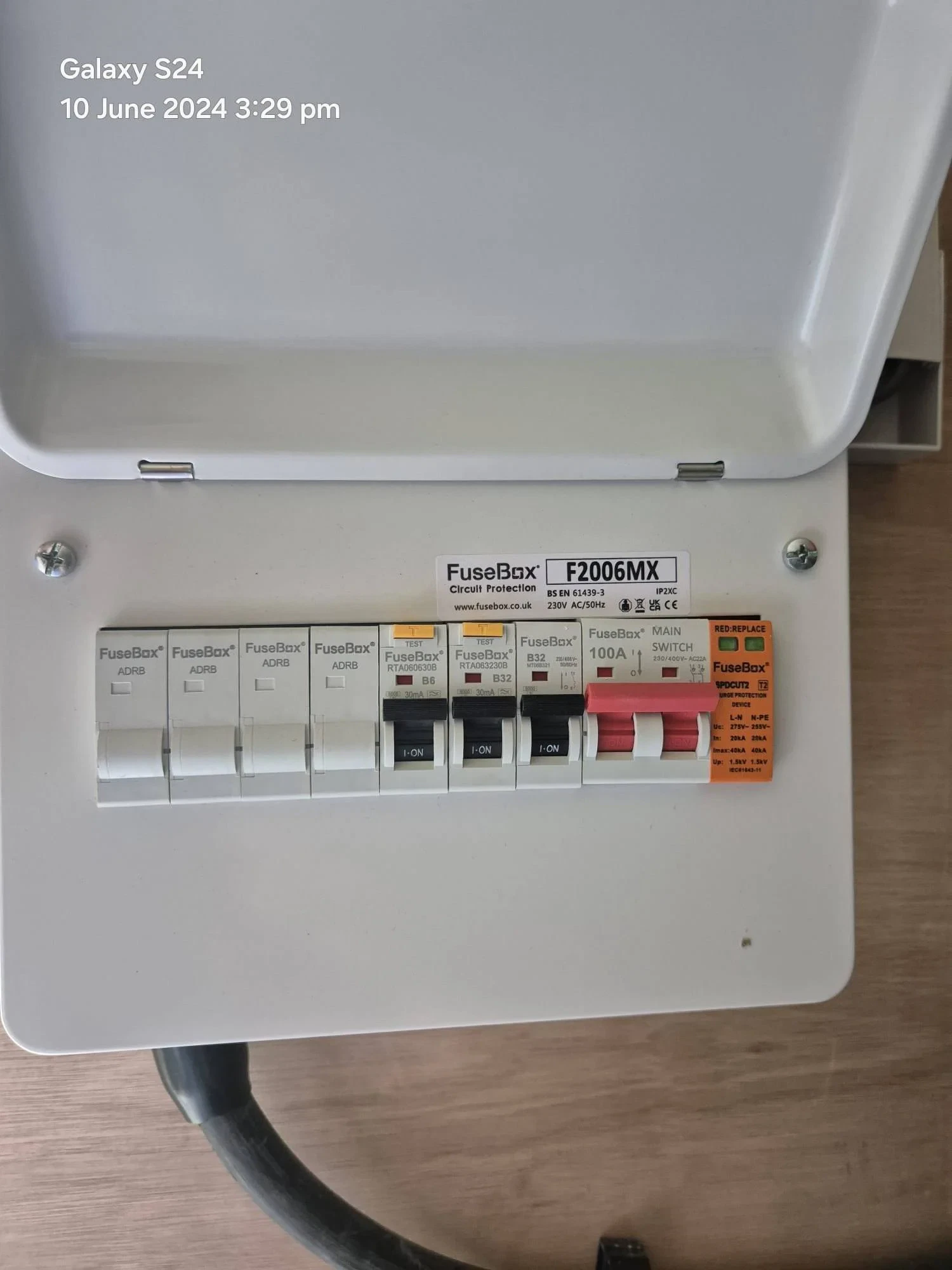 An open FuseBox consumer unit showing labelled switches, including main switch, RCD, and individual circuit breakers. The unit is mounted on a light wood surface. Date and time are displayed in the top left corner.