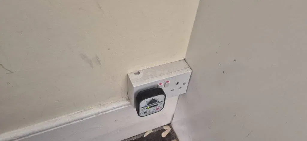 A double power socket on a wall, with a black electrical device plugged into the left socket. The socket has two red switches and is mounted near a scuffed wall and skirting board. Peeling paint is visible on the floor below.