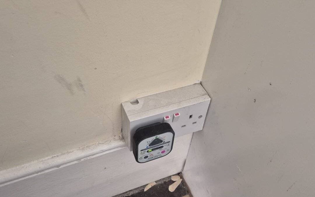 A double power socket on a wall, with a black electrical device plugged into the left socket. The socket has two red switches and is mounted near a scuffed wall and skirting board. Peeling paint is visible on the floor below.