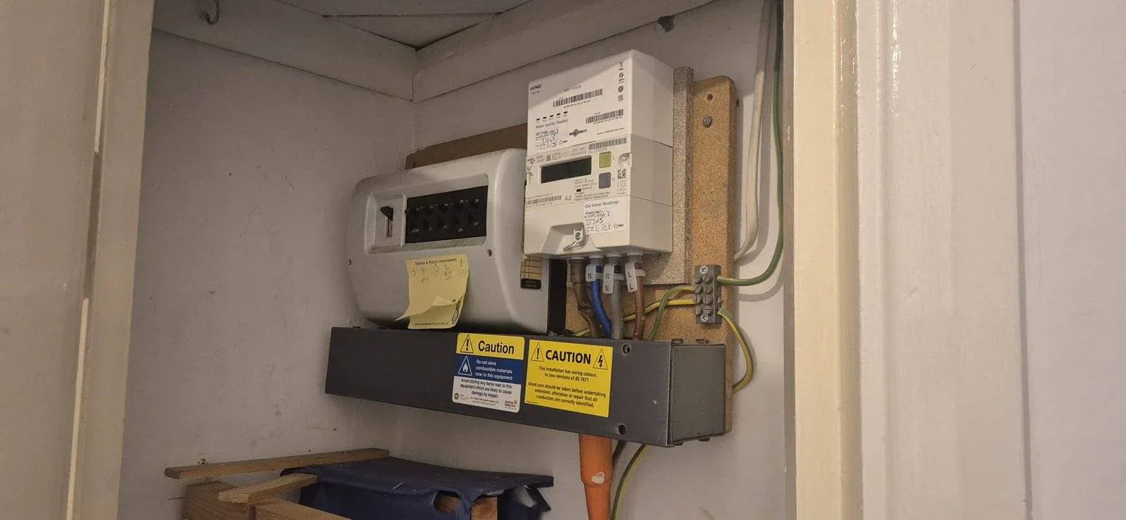 An electricity meter and fuse box are mounted on a wall inside a small cupboard. Warning and caution labels are visible, along with a handwritten sticky note on the fuse box. Various cables and pipes are connected. An electricity meter and fuse box are mounted on a wall inside a small cupboard. Warning and caution labels are visible, along with a handwritten sticky note on the fuse box. Various cables and pipes are connected.