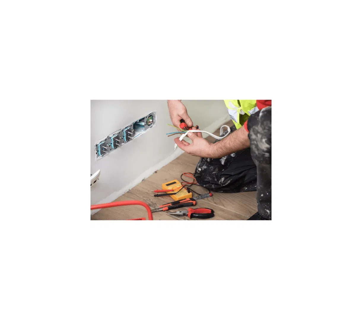A person in work clothes repairs electrical wiring next to an open wall socket, with tools such as pliers, screwdrivers, and a voltage tester on the floor—just another day for Buildspark, known for fair electrician prices in SW London.