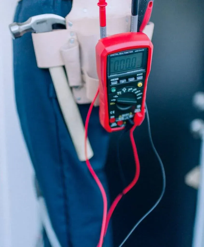 A digital multimeter with red and black probes is clipped to a tool belt worn by a person in blue trousers. A hammer and other tools are also visible in the belt.