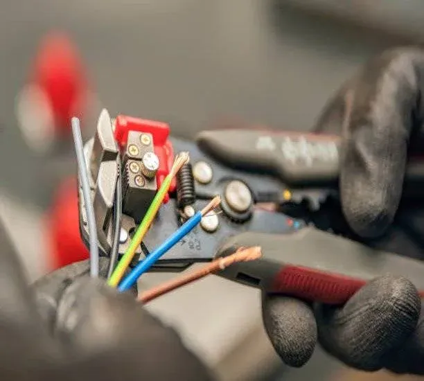 A person wearing black gloves uses a wire stripping tool to strip insulation from several coloured electrical wires, exposing the copper conductors.