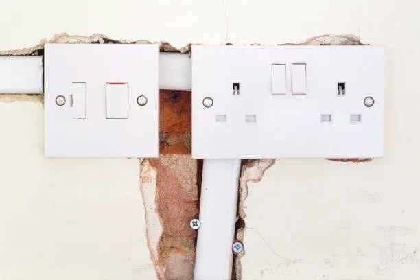 Two white electrical switch and socket plates mounted on a damaged wall, showing exposed brickwork and electrical conduit beneath the plaster.