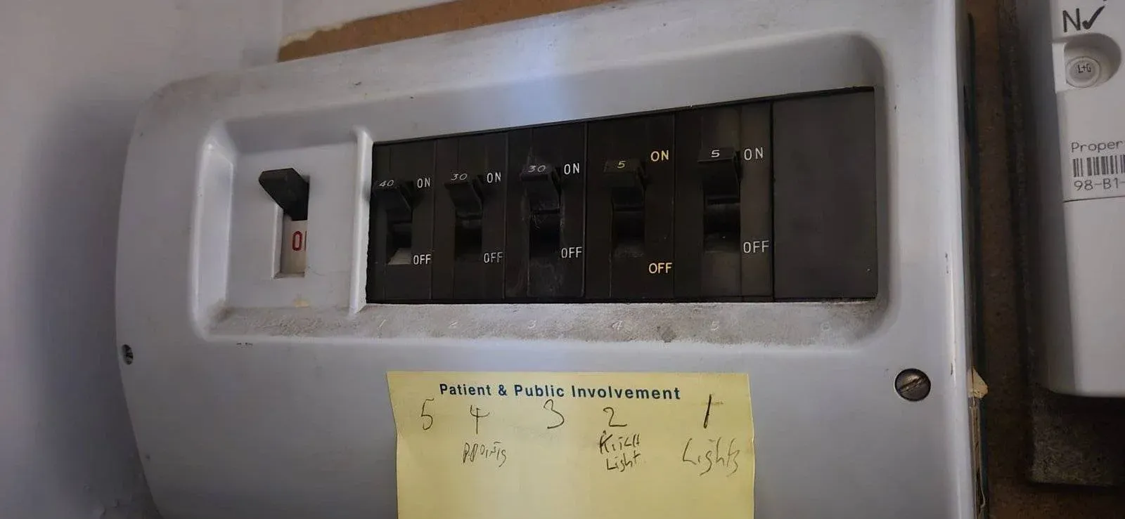 A close-up of a switchboard with six black switches, some labelled ON and OFF. A yellow sticky note below has handwritten labels and numbers for each switch, including Admin, Kitchen Light, and Lights. A close-up of a switchboard with six black switches, some labelled ON and OFF. A yellow sticky note below has handwritten labels and numbers for each switch, including Admin, Kitchen Light, and Lights.