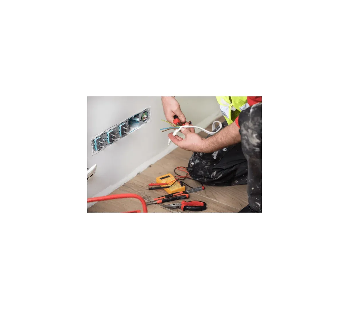 A person in work clothes repairs electrical wiring next to an open wall socket, with tools such as pliers, screwdrivers, and a voltage tester on the floor—just another day for Buildspark, known for fair electrician prices in SW London.