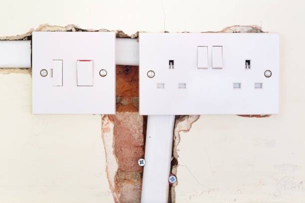 Two wall-mounted white electrical sockets and switches are shown; the plaster around them is broken, revealing red bricks and wiring beneath the surface—ideal for inspection by a certified rewiring electrician.