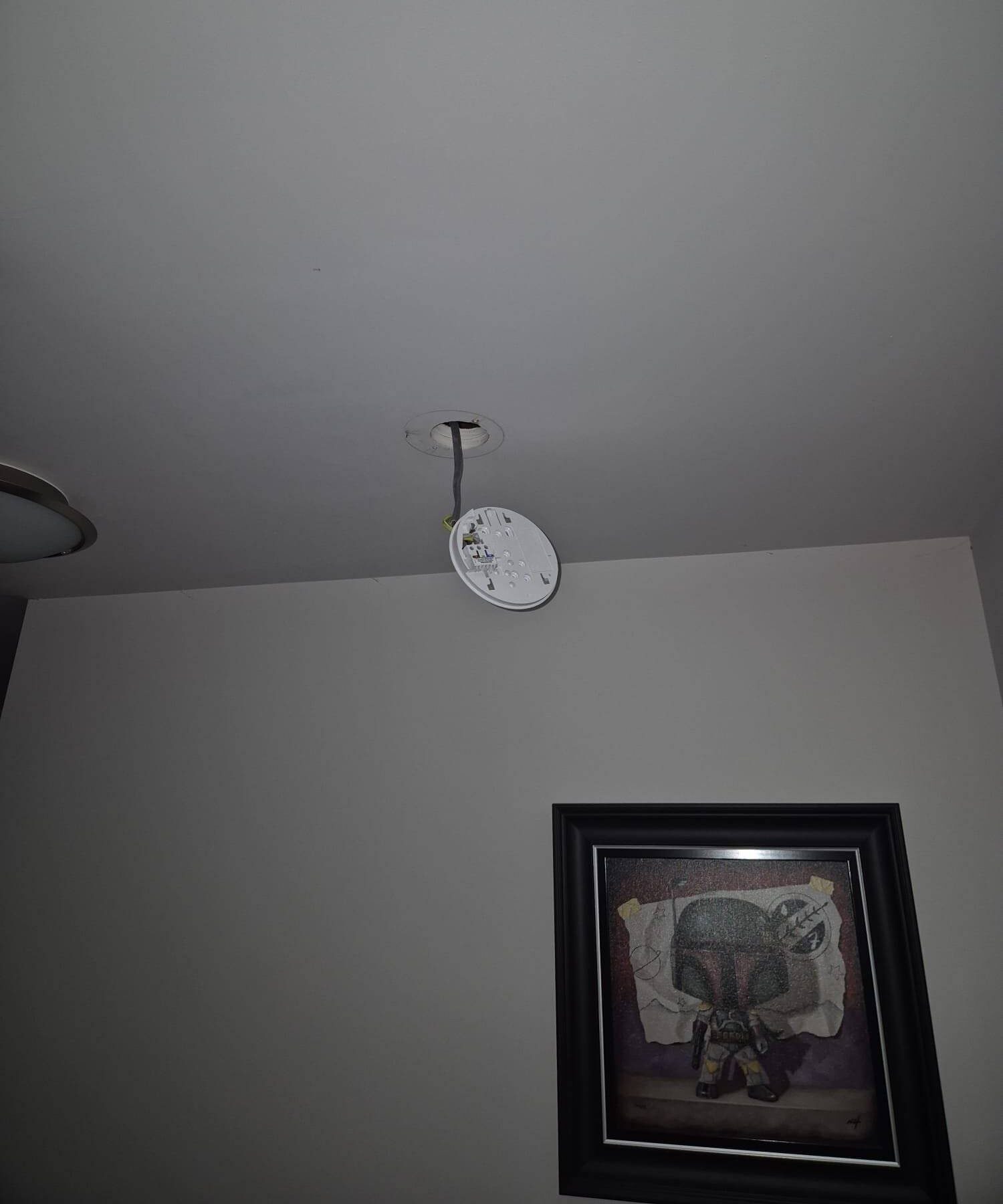 A white ceiling-mounted smoke detector hangs loose by its wires. The surrounding ceiling and wall are plain.