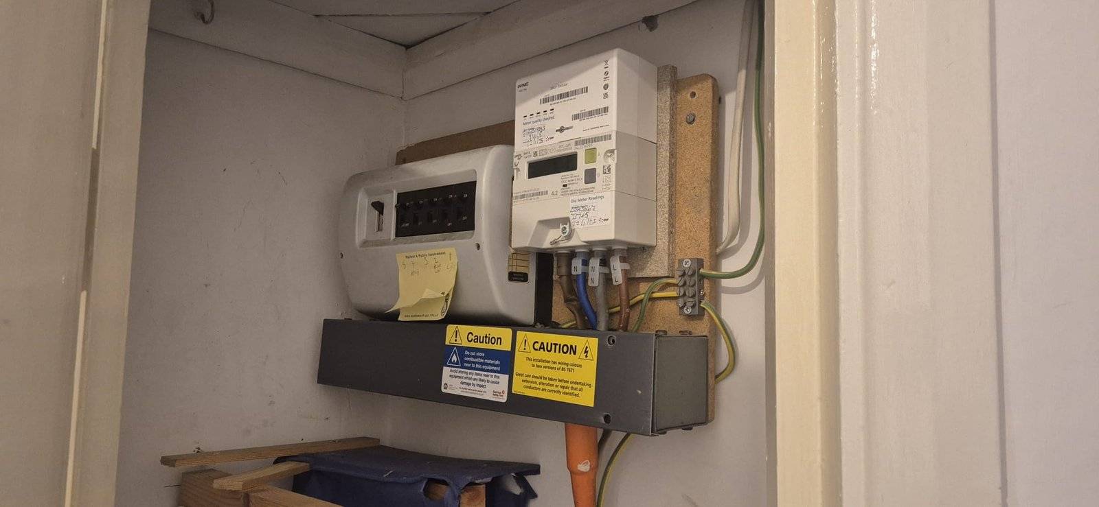 An electricity meter and fuse box are mounted on a wall inside a small cupboard. Warning and caution labels are visible, along with a handwritten sticky note on the fuse box. Various cables and pipes are connected.