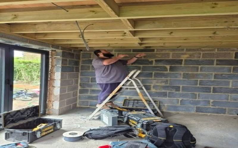 House Rewiring Services Cheam SM2 | BuildSpark Renovations An electrician standing on a stepladder installing electrical cables through timber ceiling joists in a property renovation structure, referenced as Full-Rewiring-rotated.jpg.