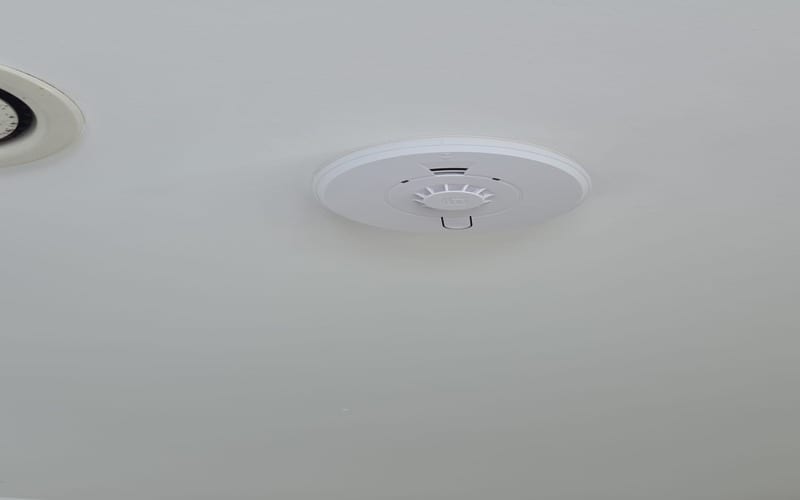 Heat Alarm Installation Cheam SM2 | BuildSpark Electricians A close-up view of a recently installed white, mains-powered fire safety alarm on a residential ceiling, referenced as Heat alarm installed cheam sm2.jpeg. The device features a modern design and is part of a certified interlinked smoke and heat detection system.